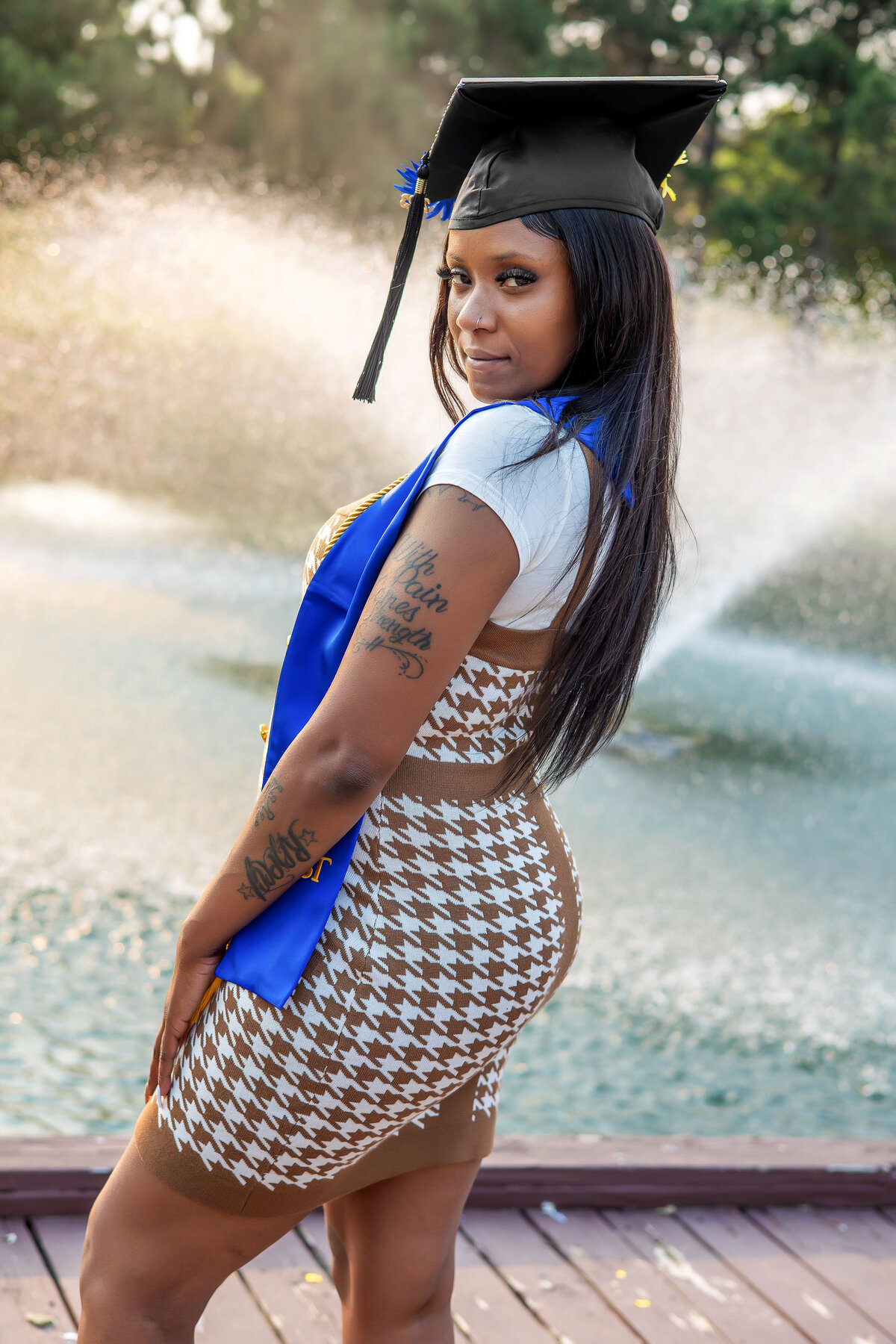 College graduate holding graduation gown, shash, and honors with back turned in front of a bridge