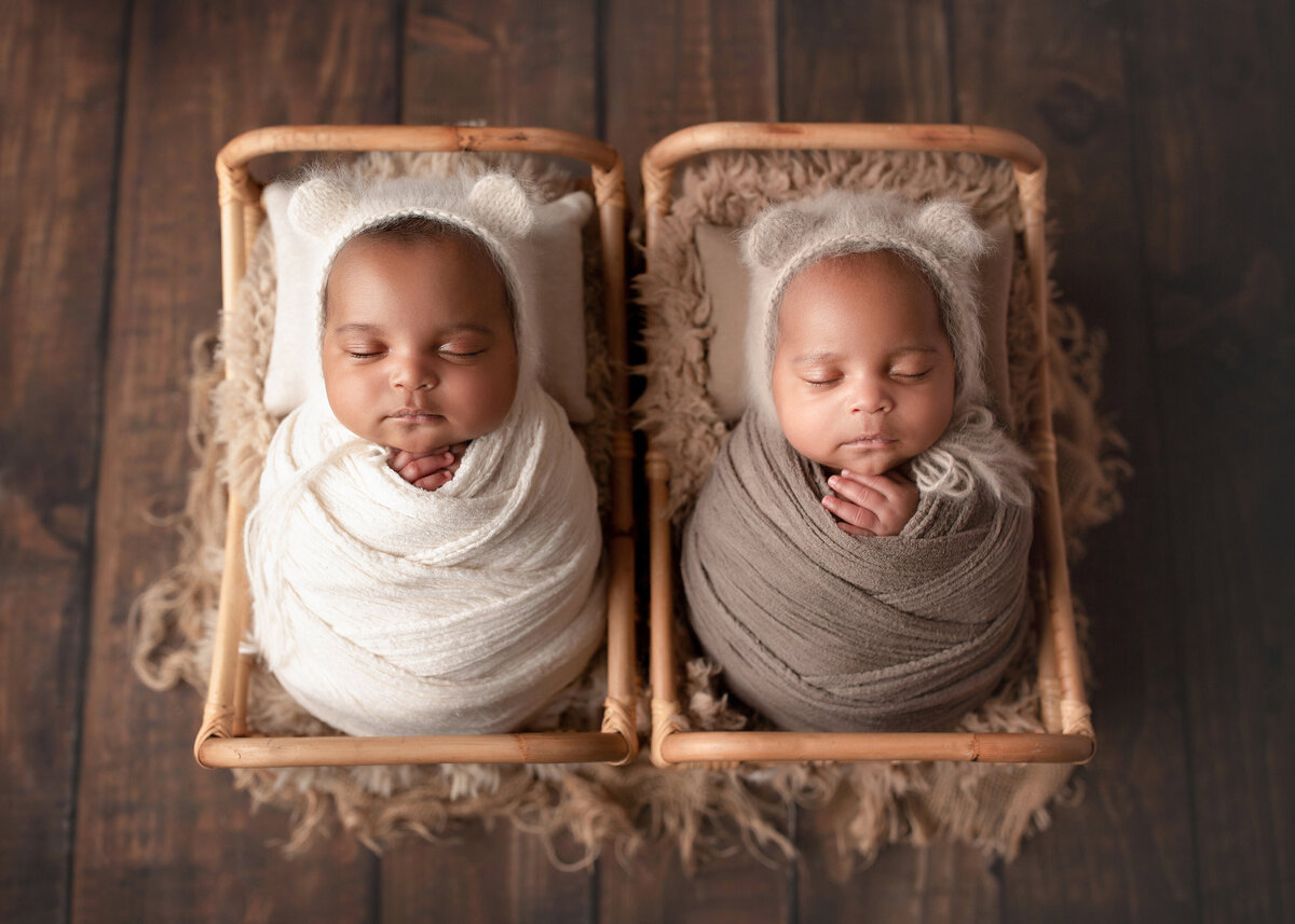 Twin baby boys sleeping in side-by-side newborn miniature cribs for newborn photo session. One twin is wrapped in white knit swaddle, the other in a taupe knit swaddle. Both boys are wearing fuzzy teddy bear hats and their hands are peeking out of the swaddle.