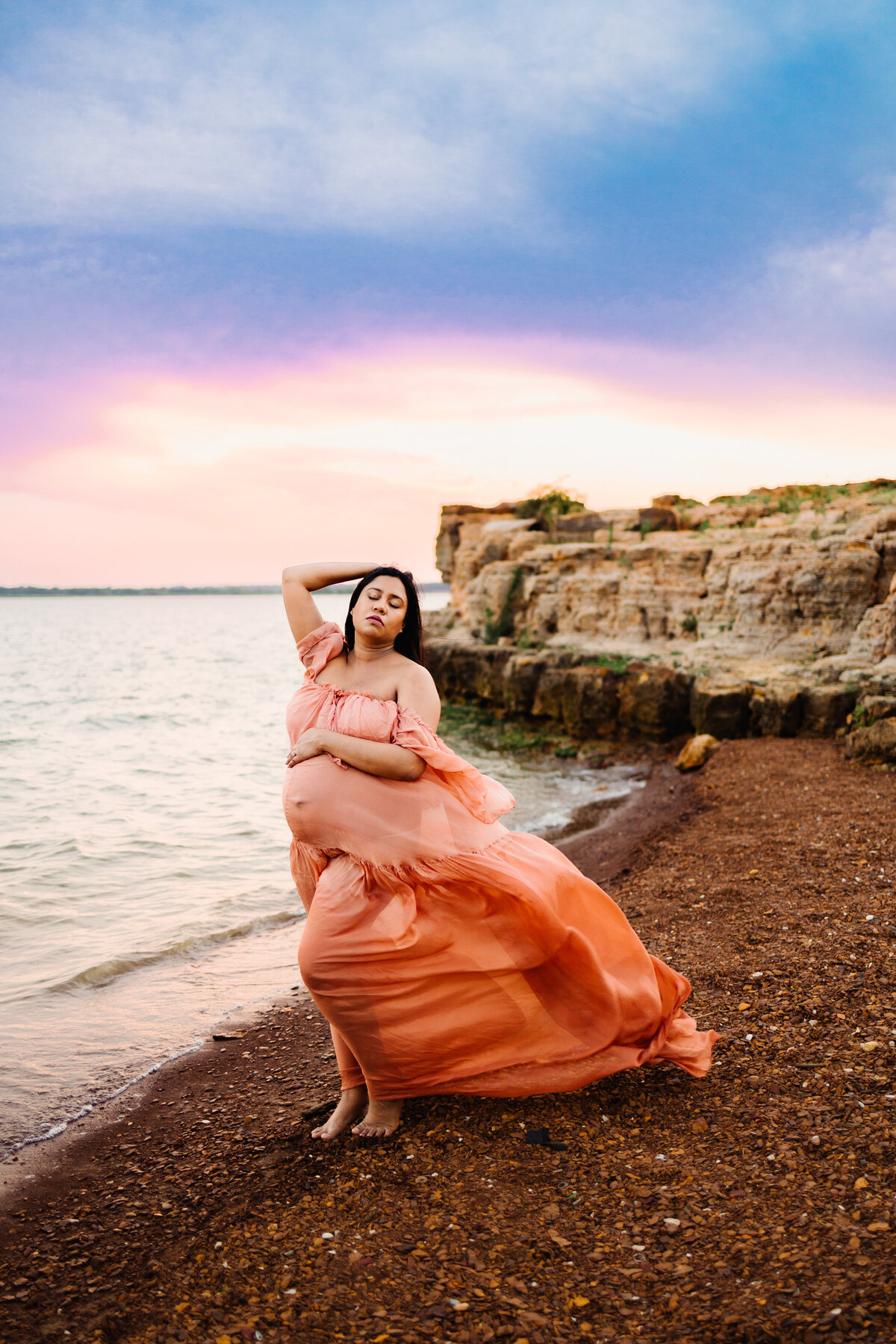 A serene maternity photo of a pregnant woman in an orange dress standing by the sea. With her hand on her head and eyes closed, she embodies tranquility and grace against a backdrop of sand dunes and a clear blue sky.