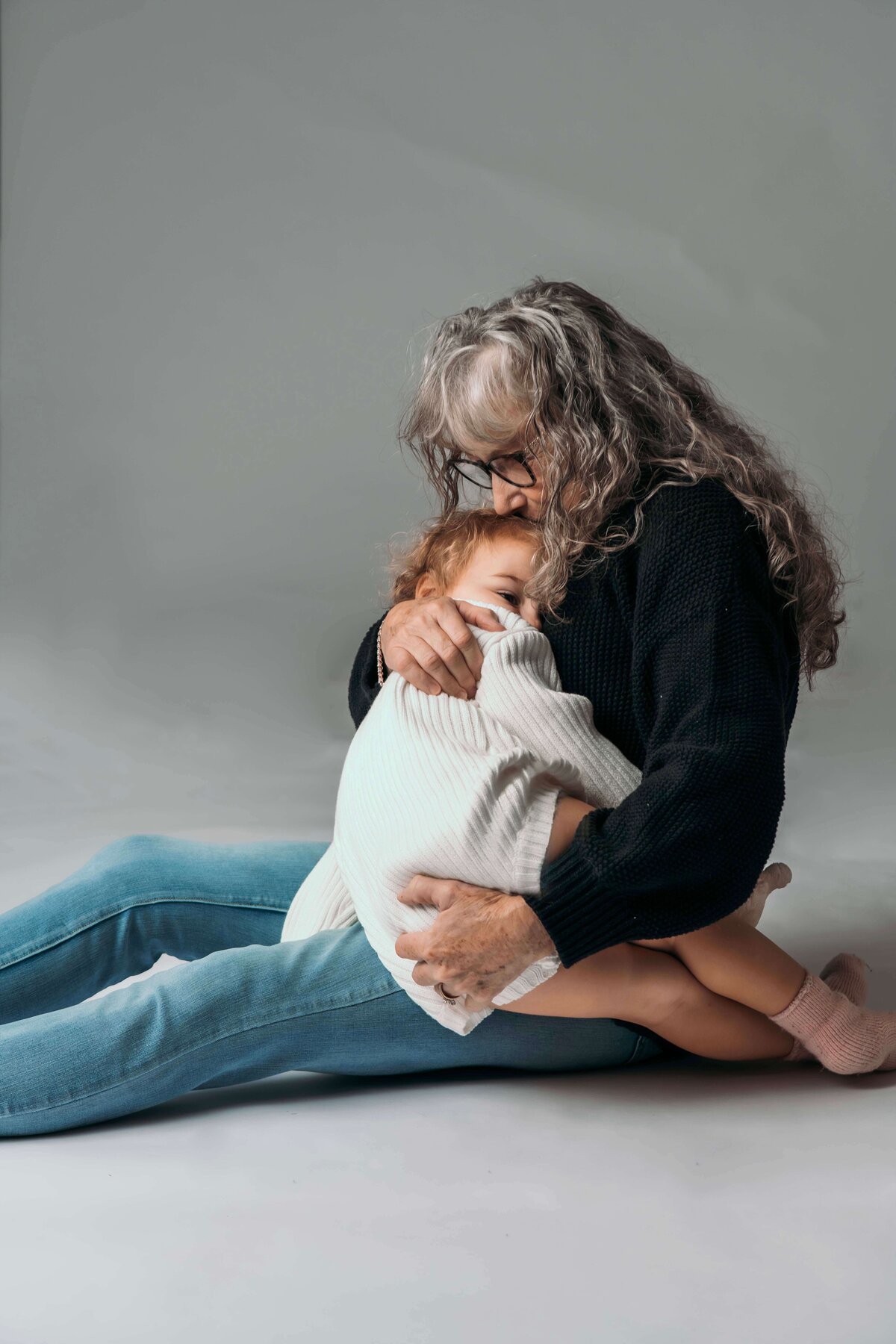 A grandmother in a cozy outfit of blue jeans, a black sweater, and grey socks sits on the floor of Mobile, AL Motherhood Photographer’s studio, embracing her 2-year-old granddaughter, who is wearing an oversized white sweater onesie. The intimate portrait captures their warm and loving connection.