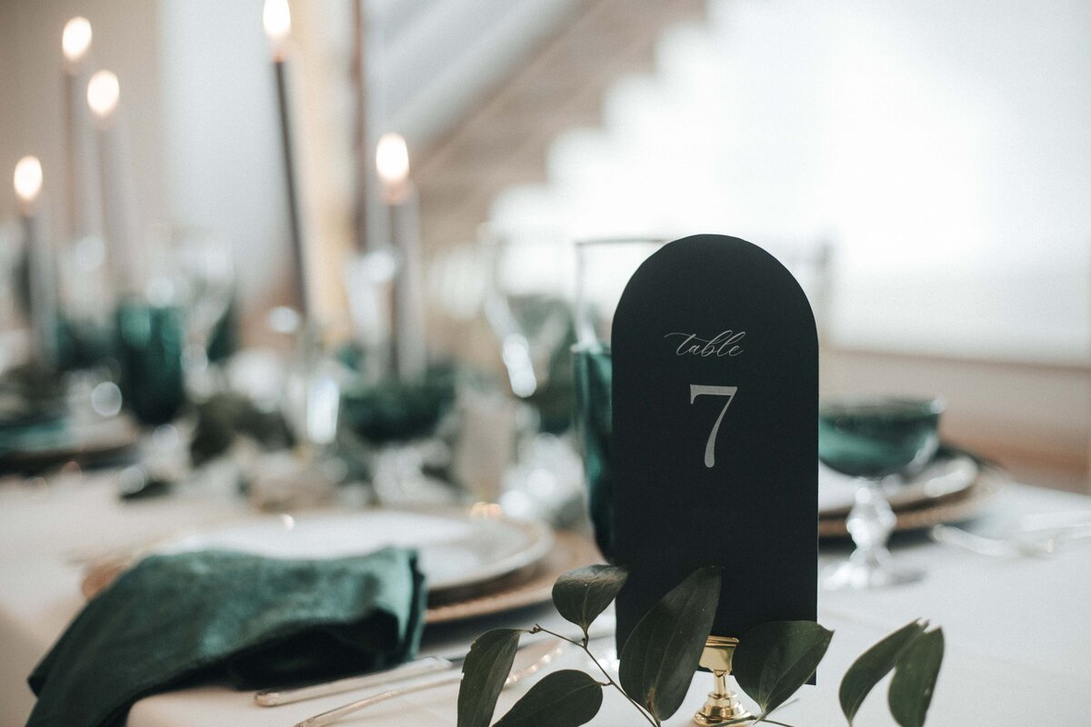 Rounded dark green place card with white cursive font on gold place card holder set atop a wedding reception table.