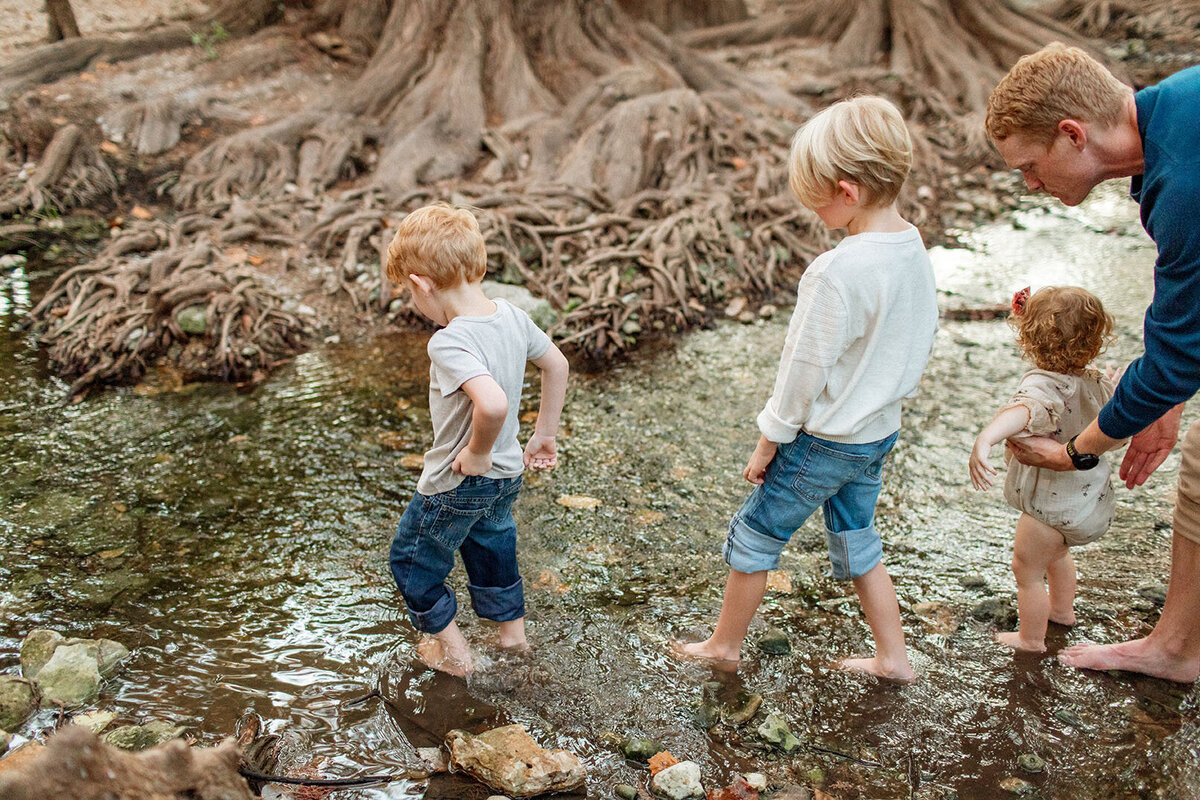 Two children standing in a river with their jeans rolled up, photographed by Sara James, a San Antonio family photographer.