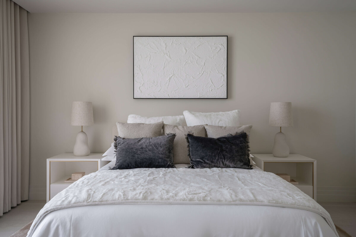 Light and airy guest room featuring a white bed, cream colored bench, and white side tables atop a soft tan rug. Above the bed is a white art piece.