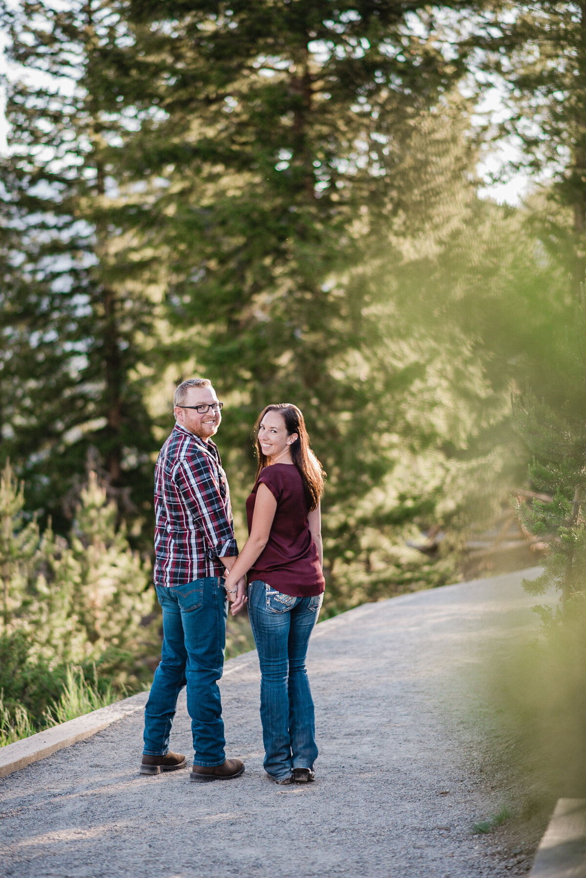 Couple walks along a dirt path on a mountain trail while holding hands and then turn halfway around and smile at the camera