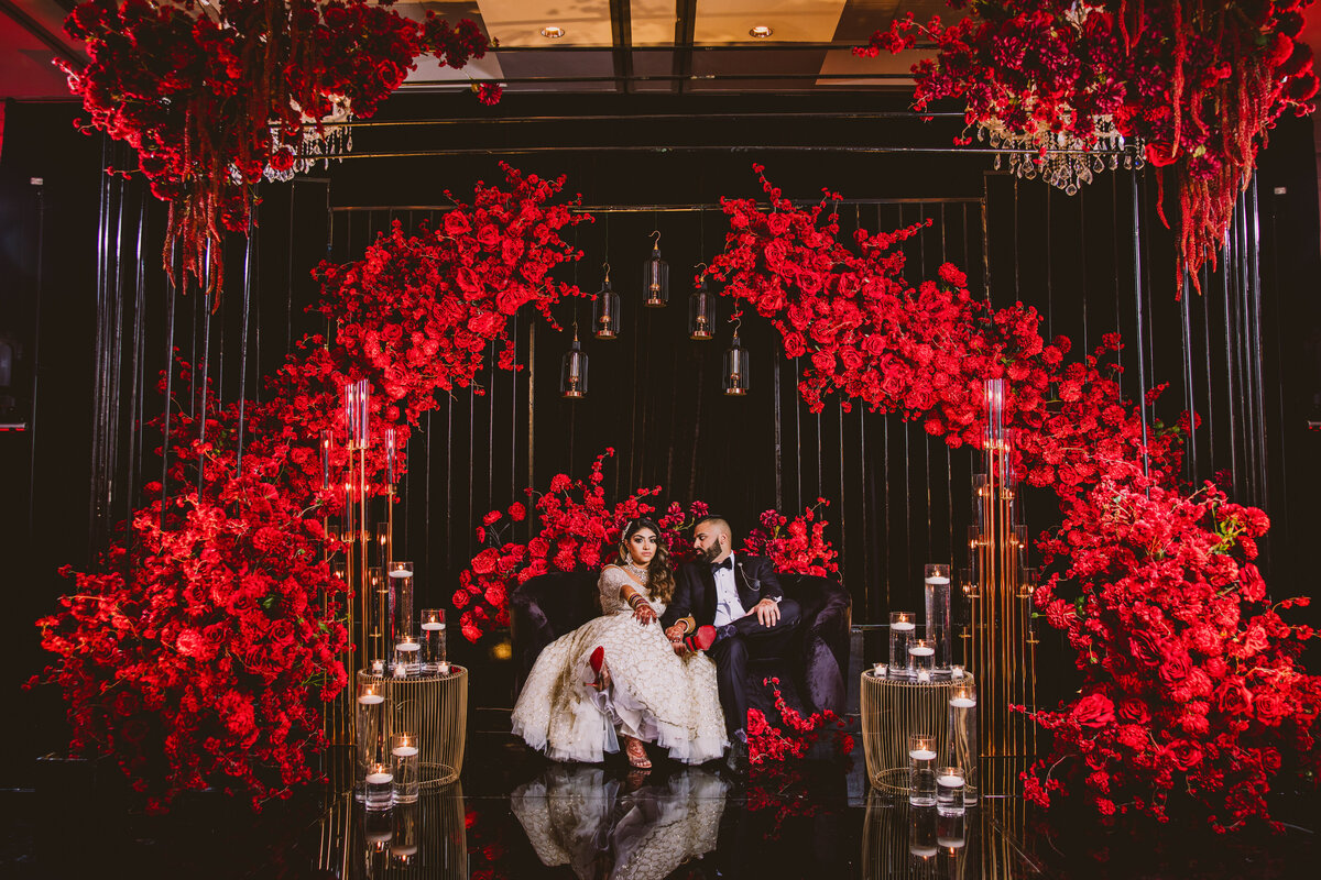 indian bride and groom sitting on a dark, black and red decorated stage at their wedding reception at addison park nj