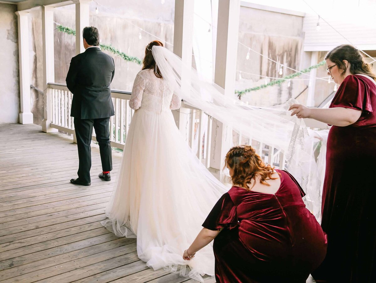 A bride stands behind a man in a black suite, facing away. Two bridesmaids in burgundy velvet dresses arrange the bride's dress and veil for a first look.