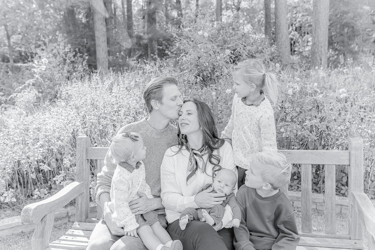 Heartwarming black and white family portrait featuring parents and their four children sharing a loving moment on a garden bench—captured by a Birmingham, AL family photographer.