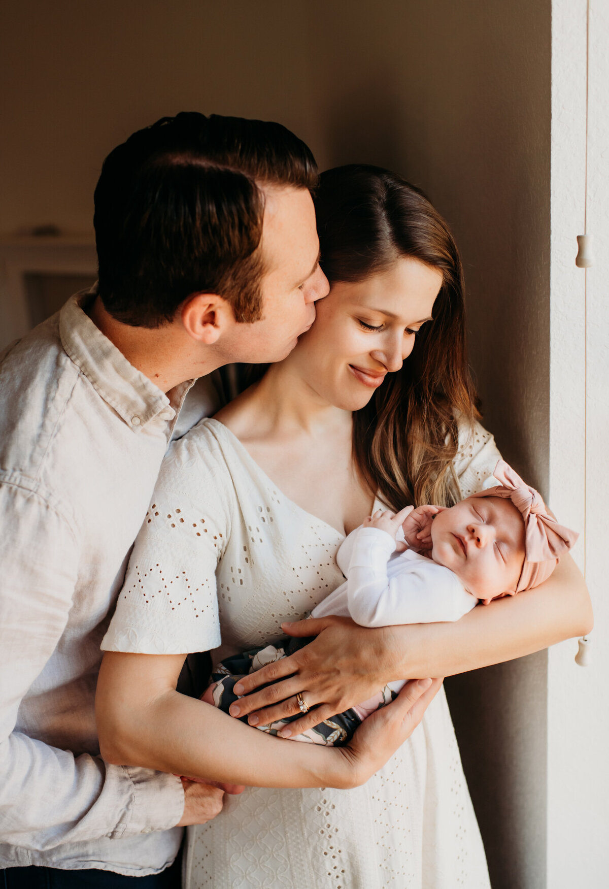 Newborn Photographer, Dad is kissing mom, who is holding baby girl by the window.