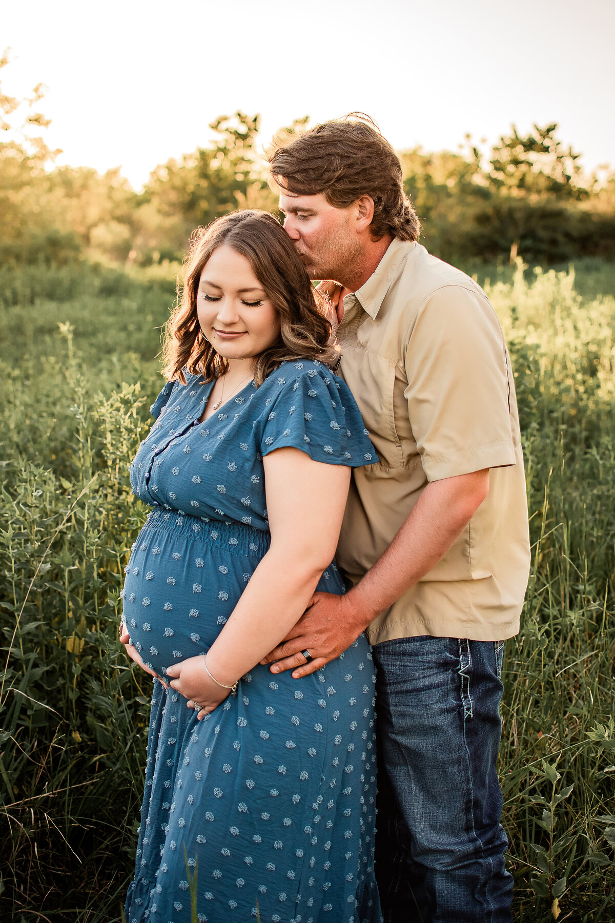 An expectant father stands behind his wife and kisses her head as she holds her belly.