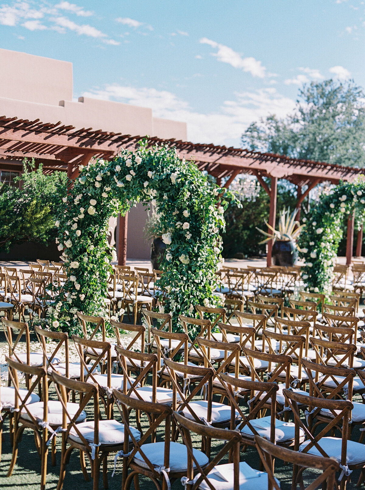 A gorgeous wedding ceremony setup featuring floral archways and stylish wooden seating under a clear blue sky. A dream setting for any couple planning their big day.