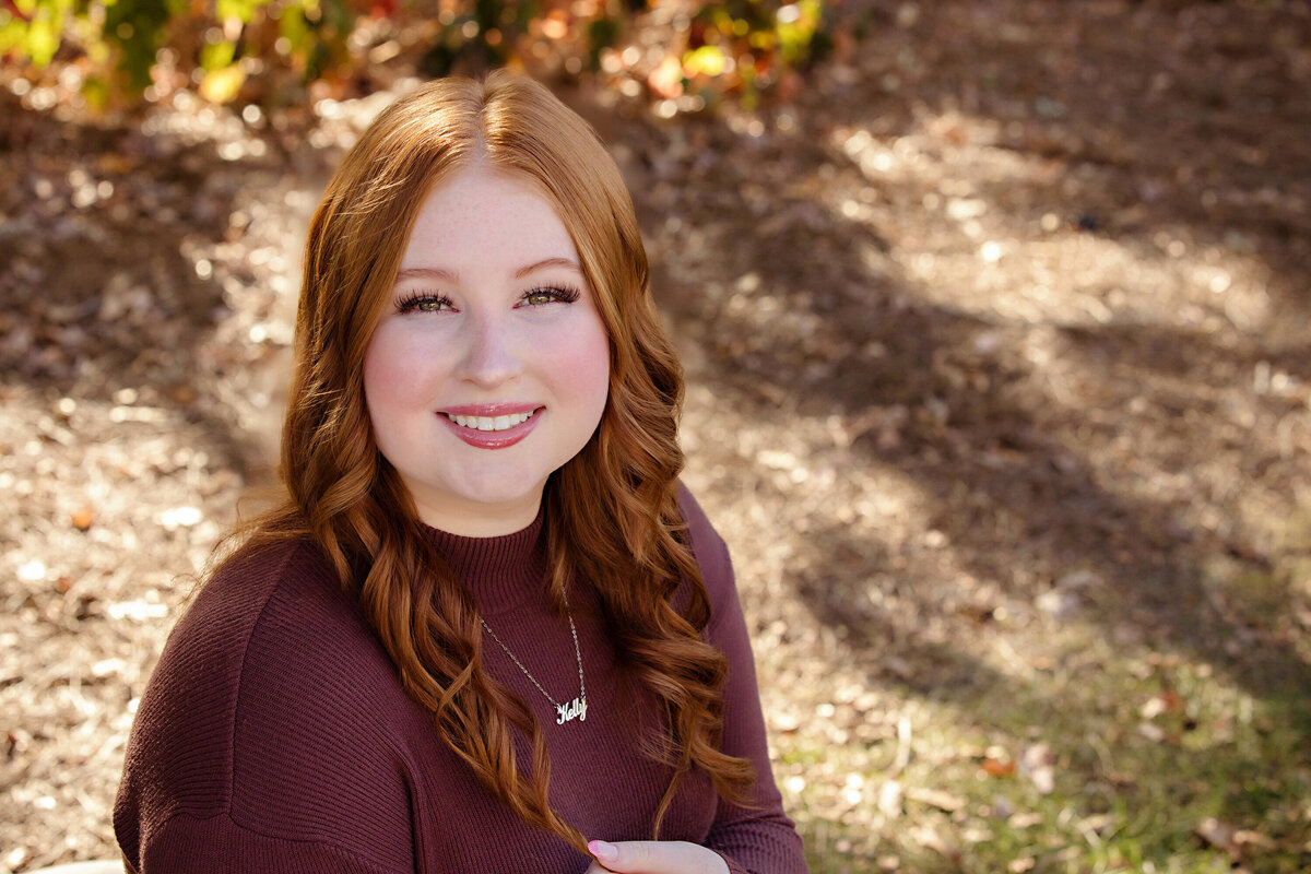 High school senior in cranberry-toned top and jeans smiling softly in open grassy space with fall light filtering through nearby trees