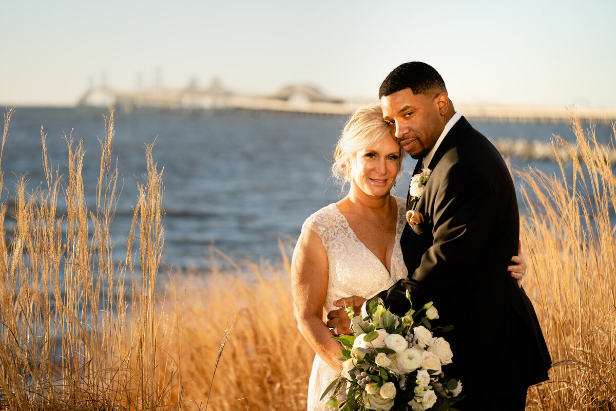 A bride and groom with their arms around each other standing in front of a lake.