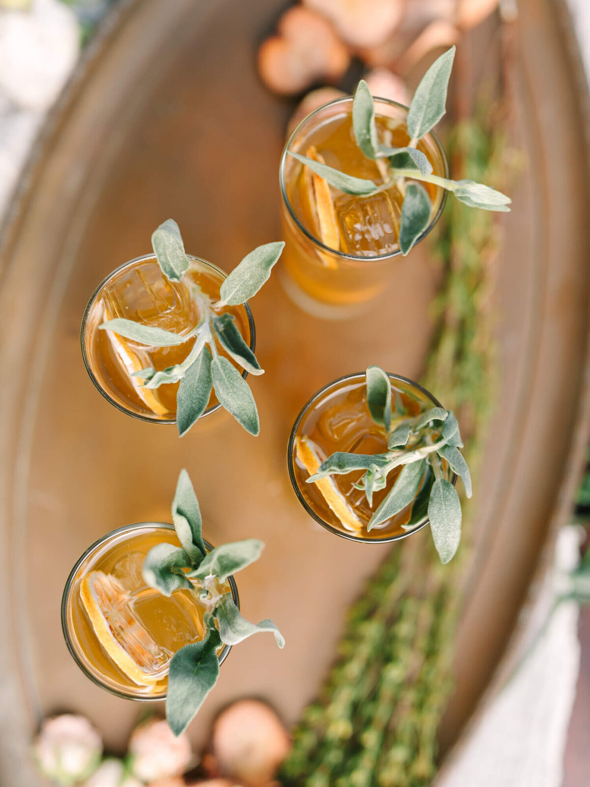 Top-down view of four amber-colored drinks in glasses garnished with fresh sage leaves on a bronze tray.