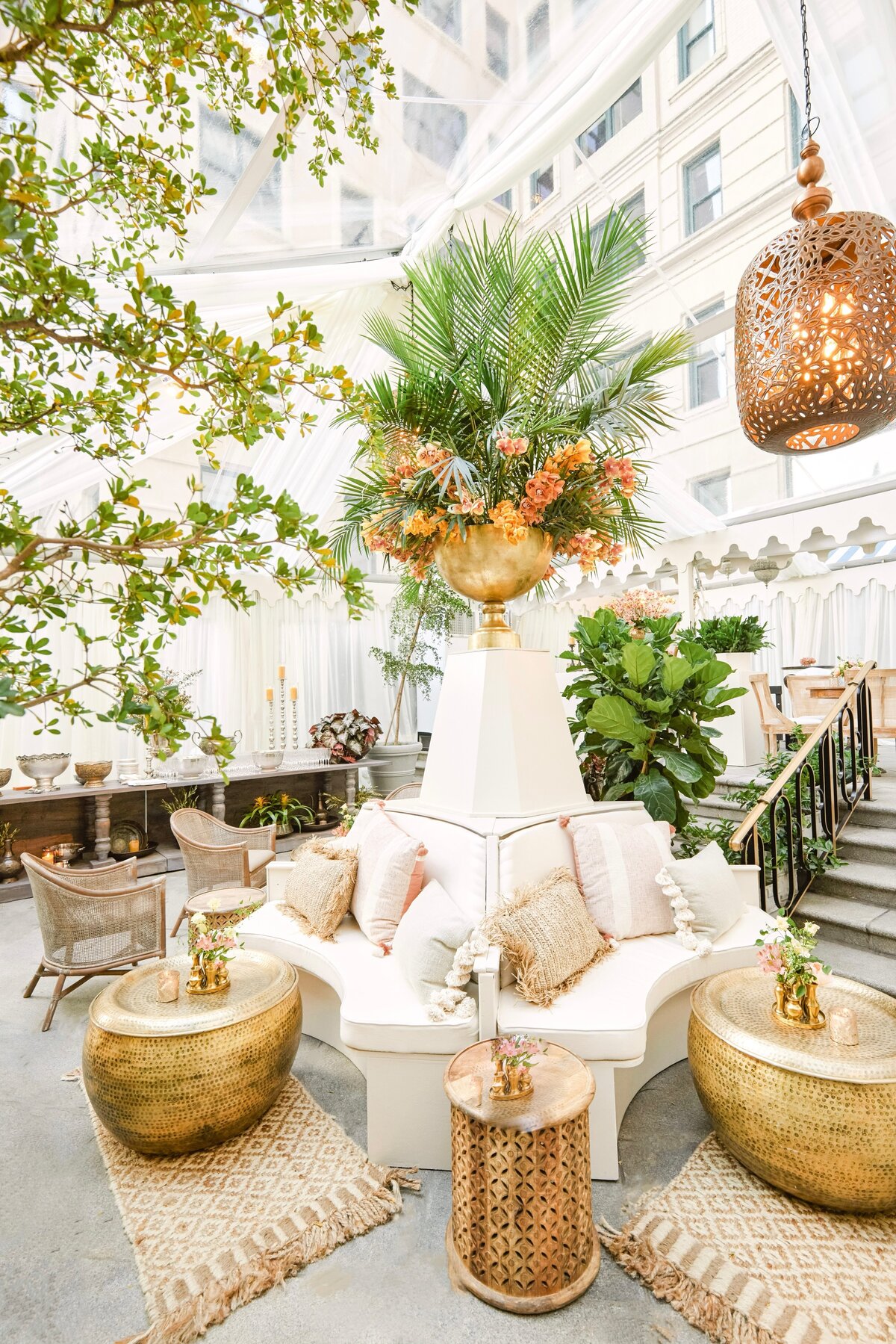 This elegant and airy room with large windows is located at The Willard Intercontinental in Washington D.C., recognized as one of the top wedding venues and hotels in the area. The photograph was taken by photographer Shawn Connell, capturing the beauty of the space. Clayton Films also provided videography services for this event. The white seating area adorned with plush cushions is complemented by gold tables, while green plants and a large floral centerpiece in a gold vase add a touch of lushness. The sophisticated decor is further enhanced by the presence of patterned rugs and hanging woven lamps, creating a truly enchanting setting.