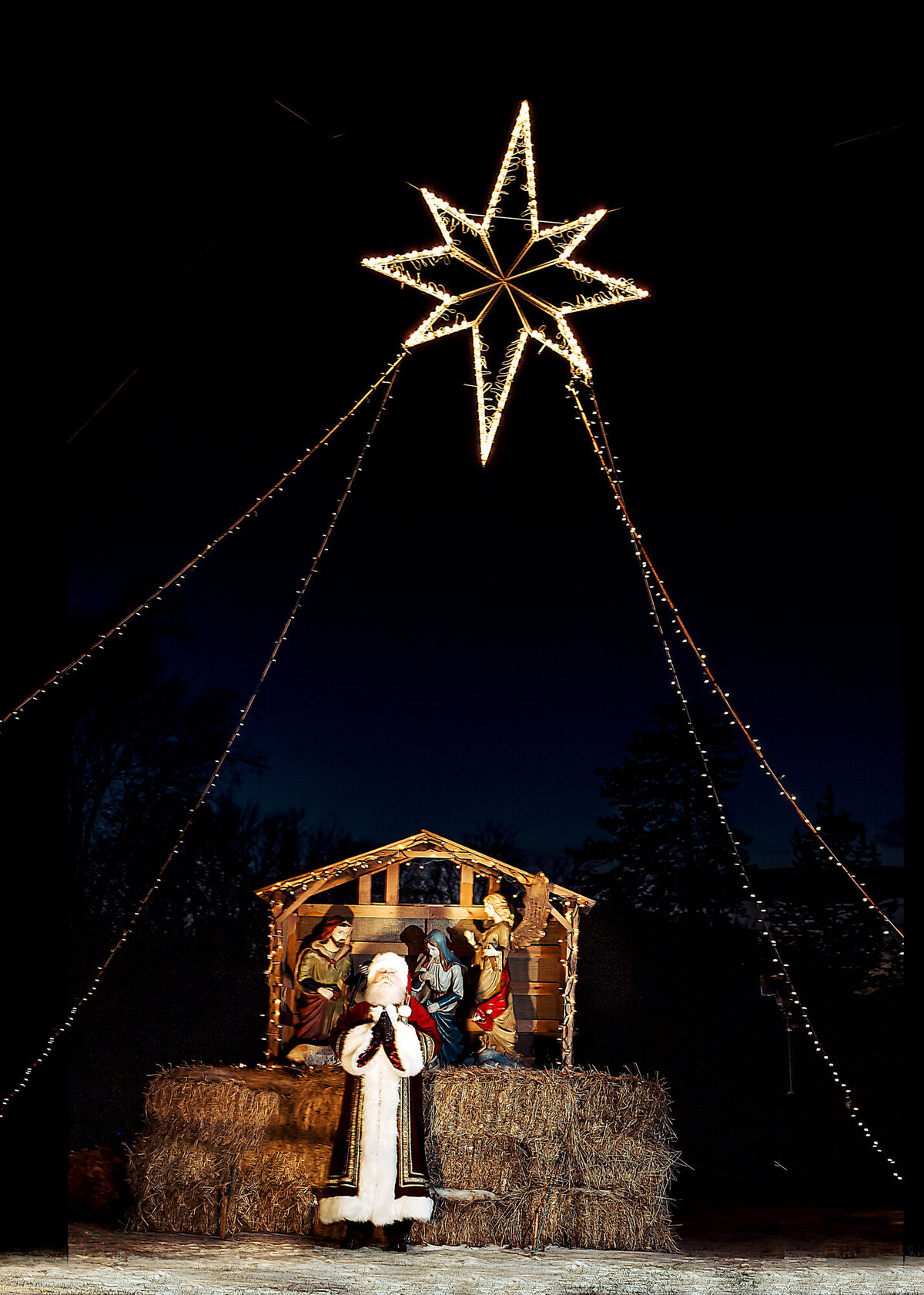 Santa Claus is standing outside in front of a manger at night with a star lit up high above him, posing for a South Shore MA branding photographer