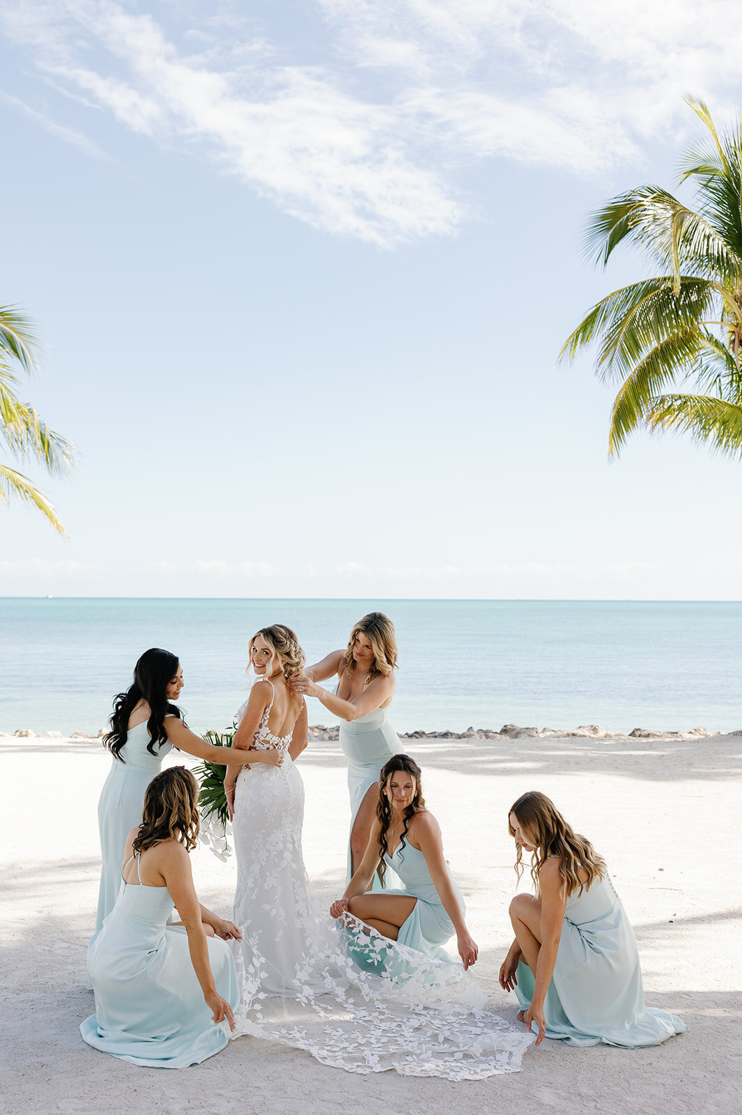 Ashley stands on a sandy beach while her bridesmaids in pastel blue dresses fix her gown.