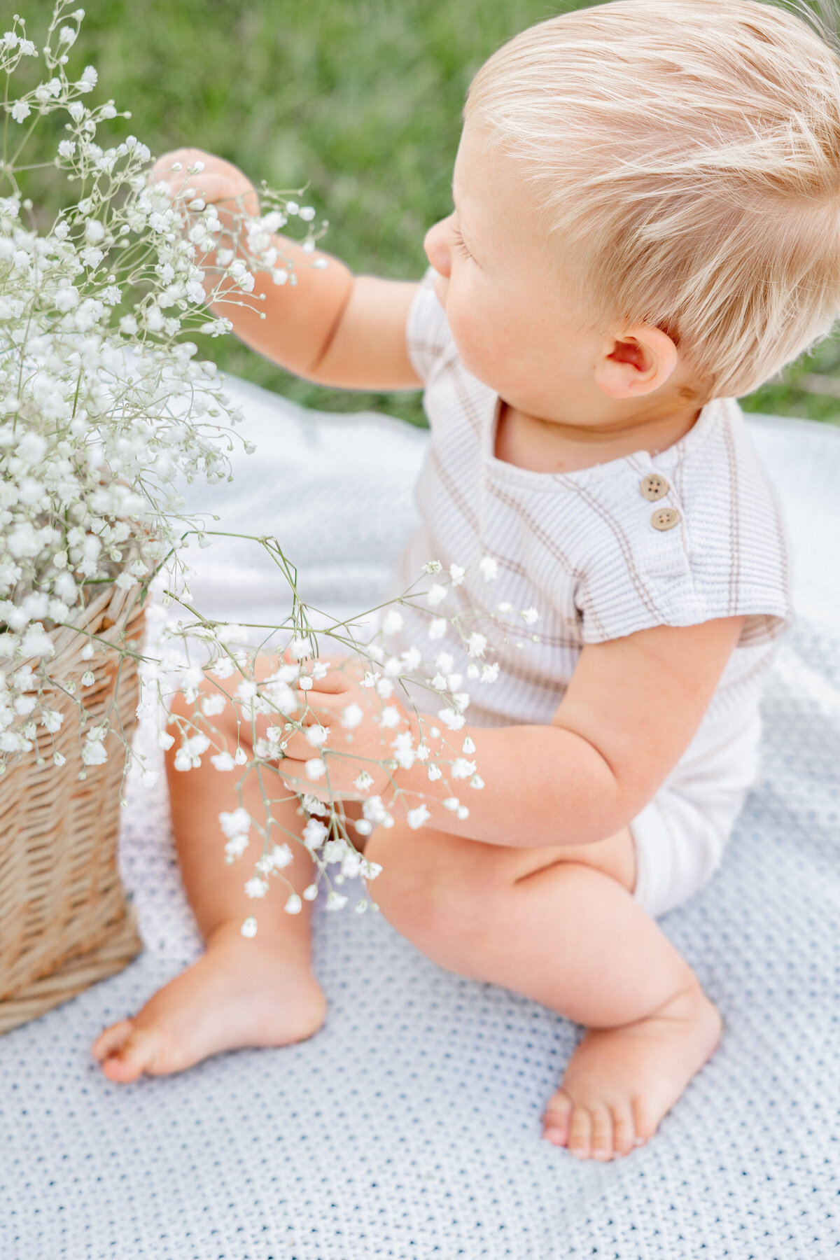 A curious baby boy reaches for delicate white wildflowers in a woven basket while sitting on a cozy blanket. Captured by Cassie Lee Photography.
