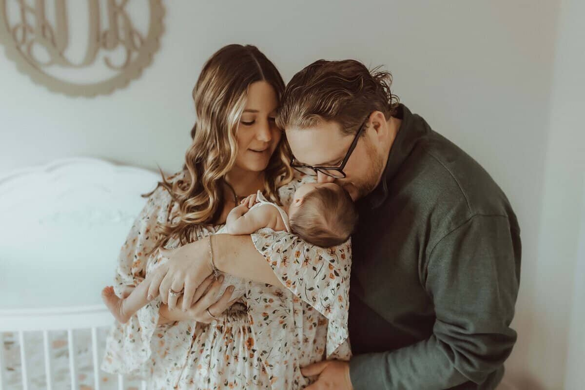 A woman holds a baby while a man leans in to kiss the baby's head; they stand together in a softly lit room.