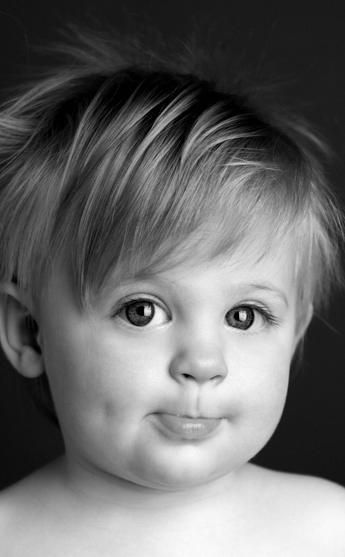 A close-up black-and-white photo of a toddler with tousled hair and a slight smile. The child's round eyes and smooth skin convey innocence and curiosity.