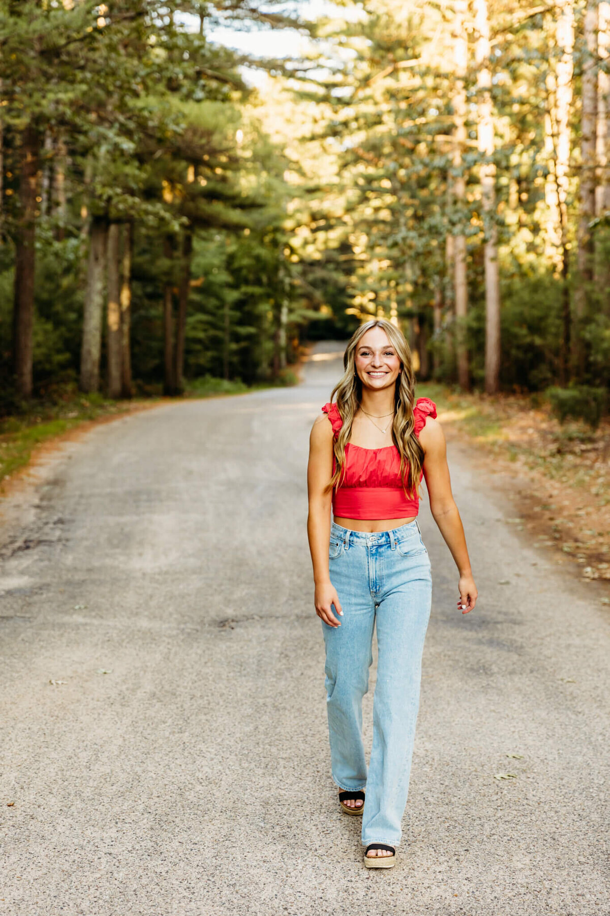 teenage girl in a red crop top, light blue jeans and sandals walking and laughing on a back road near Oshkosh