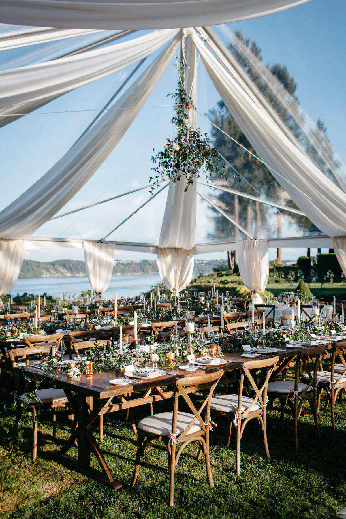 Tent wedding reception by the water with greenery covered chandeliers.