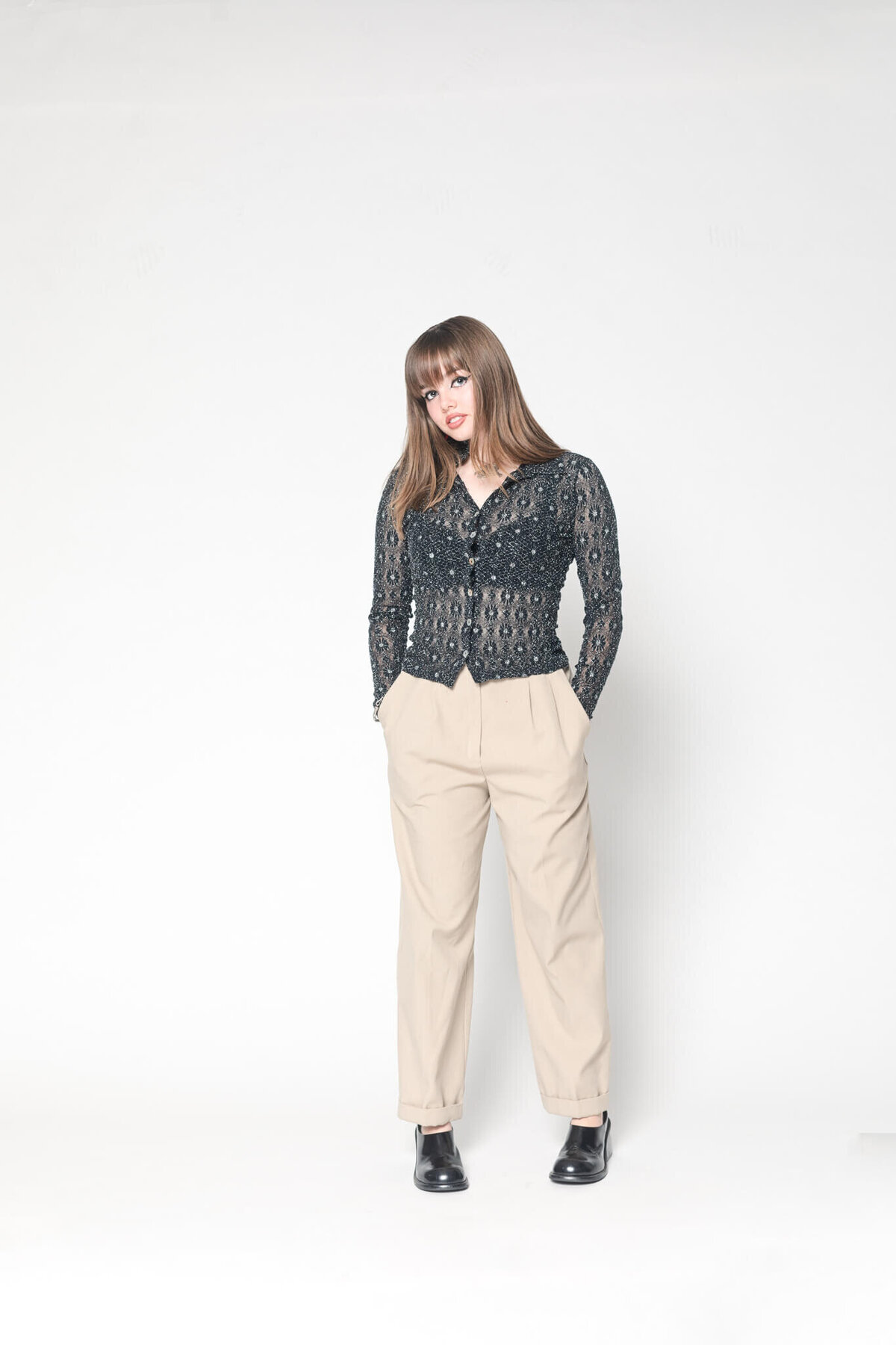 A person with long brown hair is standing against a plain white background, wearing a textured dark jacket and beige pants. They have their hands in their pockets and are looking slightly to the side.