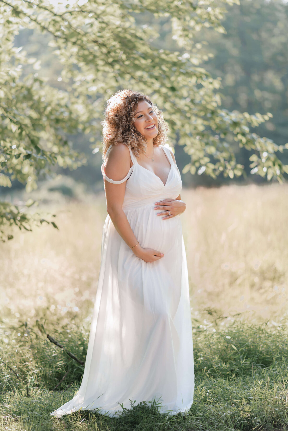 angelical mom wearing a long white maternity dress, smiling under a tree on a sunny summer day