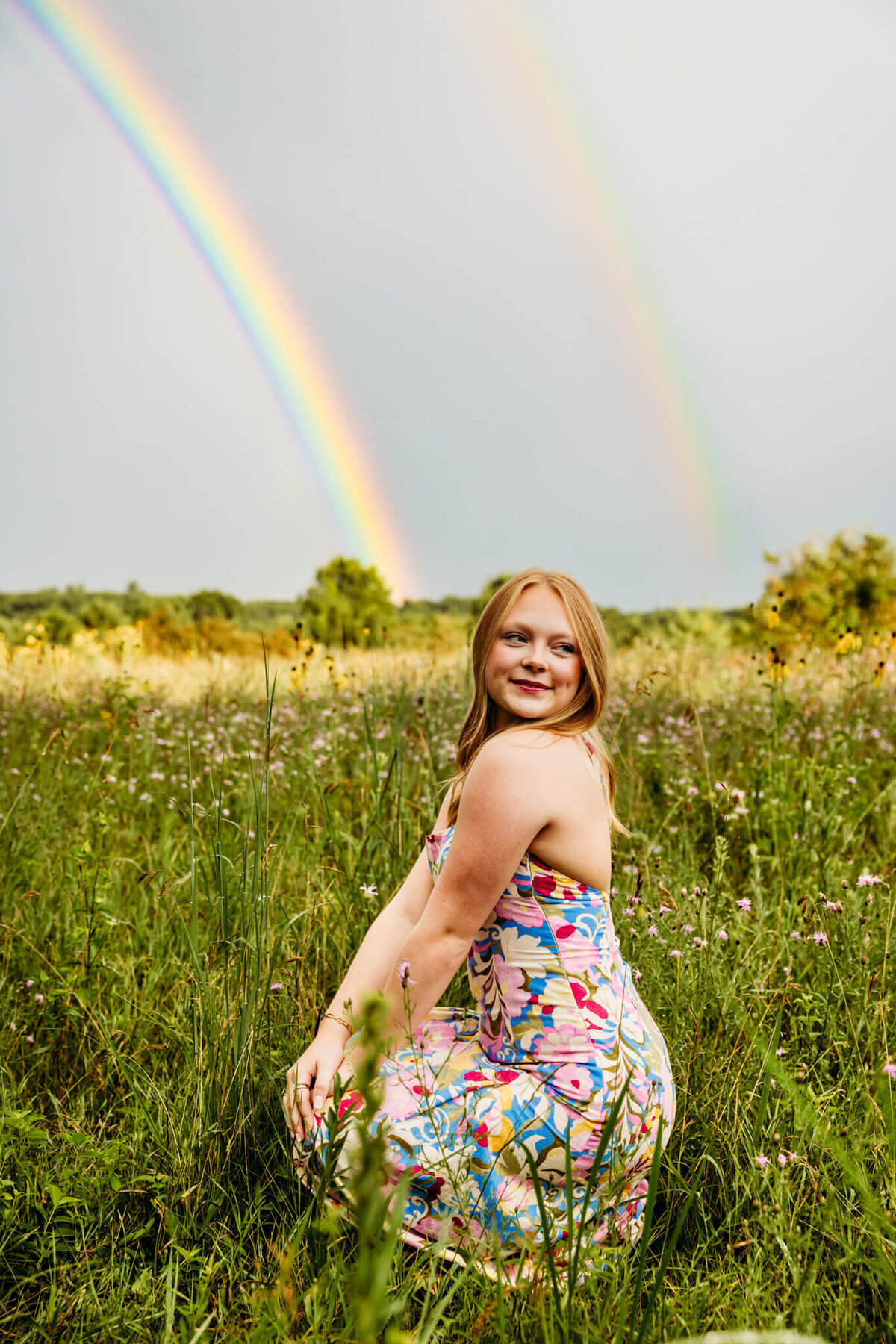 amazing photo of a teen girl in a colorful patterned dress kneeling and looking over her shoulder as two rainbows sit in the sky behind her in a field near Green Bay.