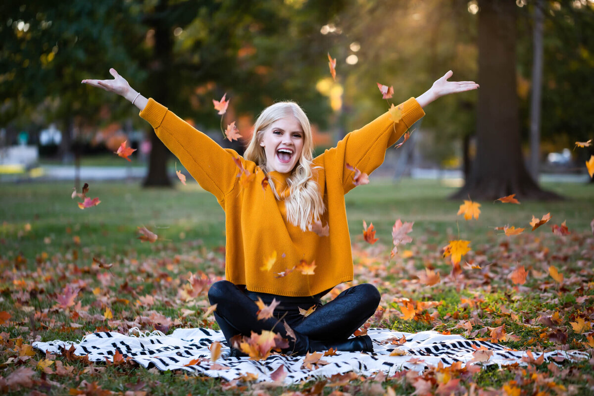 A beautiful blonde teen sitting on a black and white blanket and throwing colorful Fall leaves in the air. Captured by Springfield, MO senior photographer Dynae Levingston.