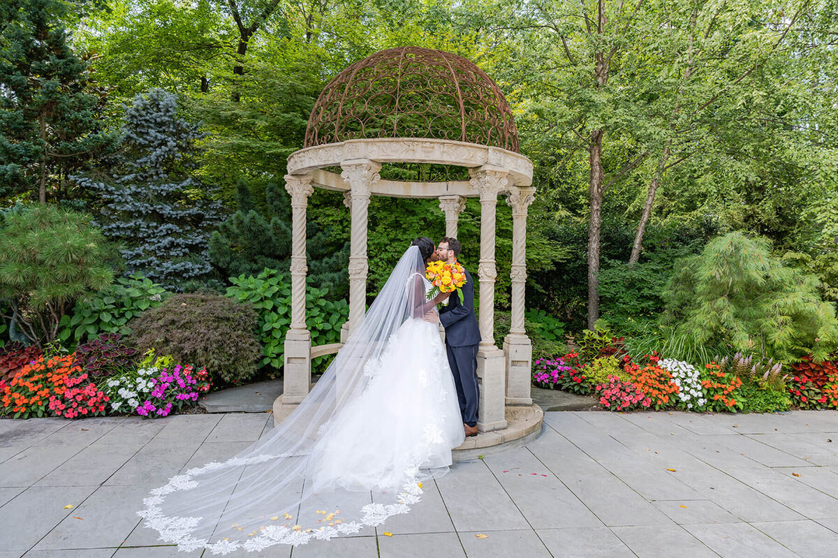 Couple sharing a romantic kiss under a stone garden gazebo surrounded by colorful flowers, captured as part of a wedding day photo timeline by Miami wedding photographer White House Wedding Photography.