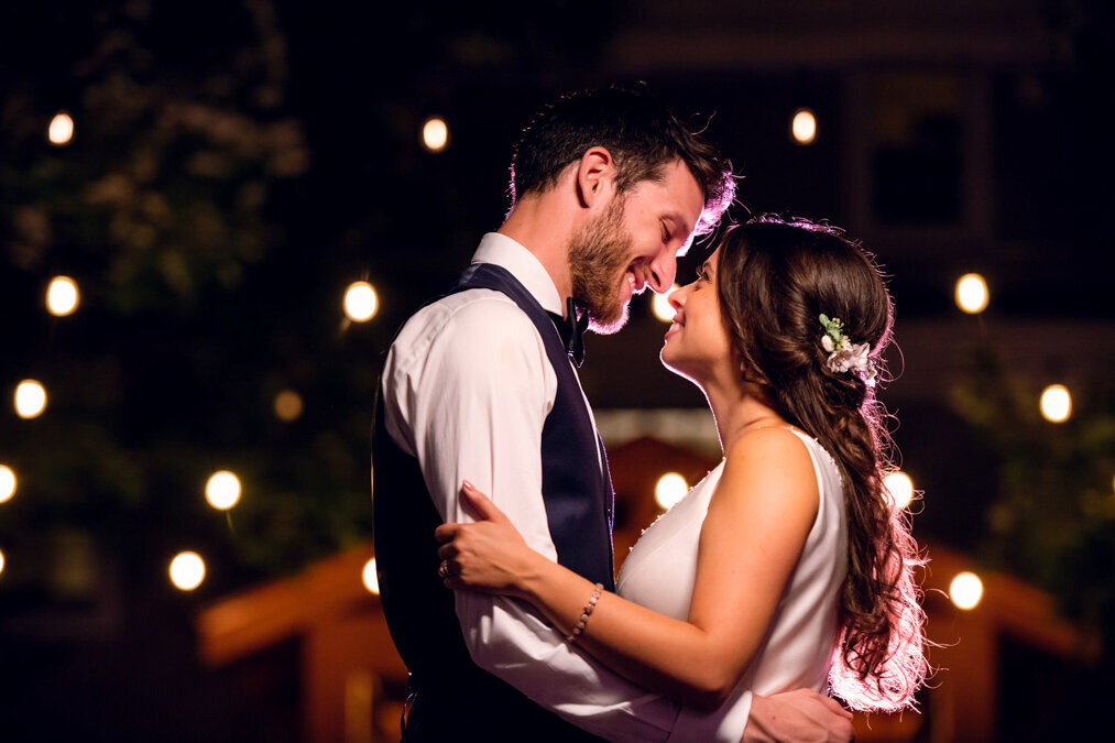 A bride and groom embrace in front of a string of lights.