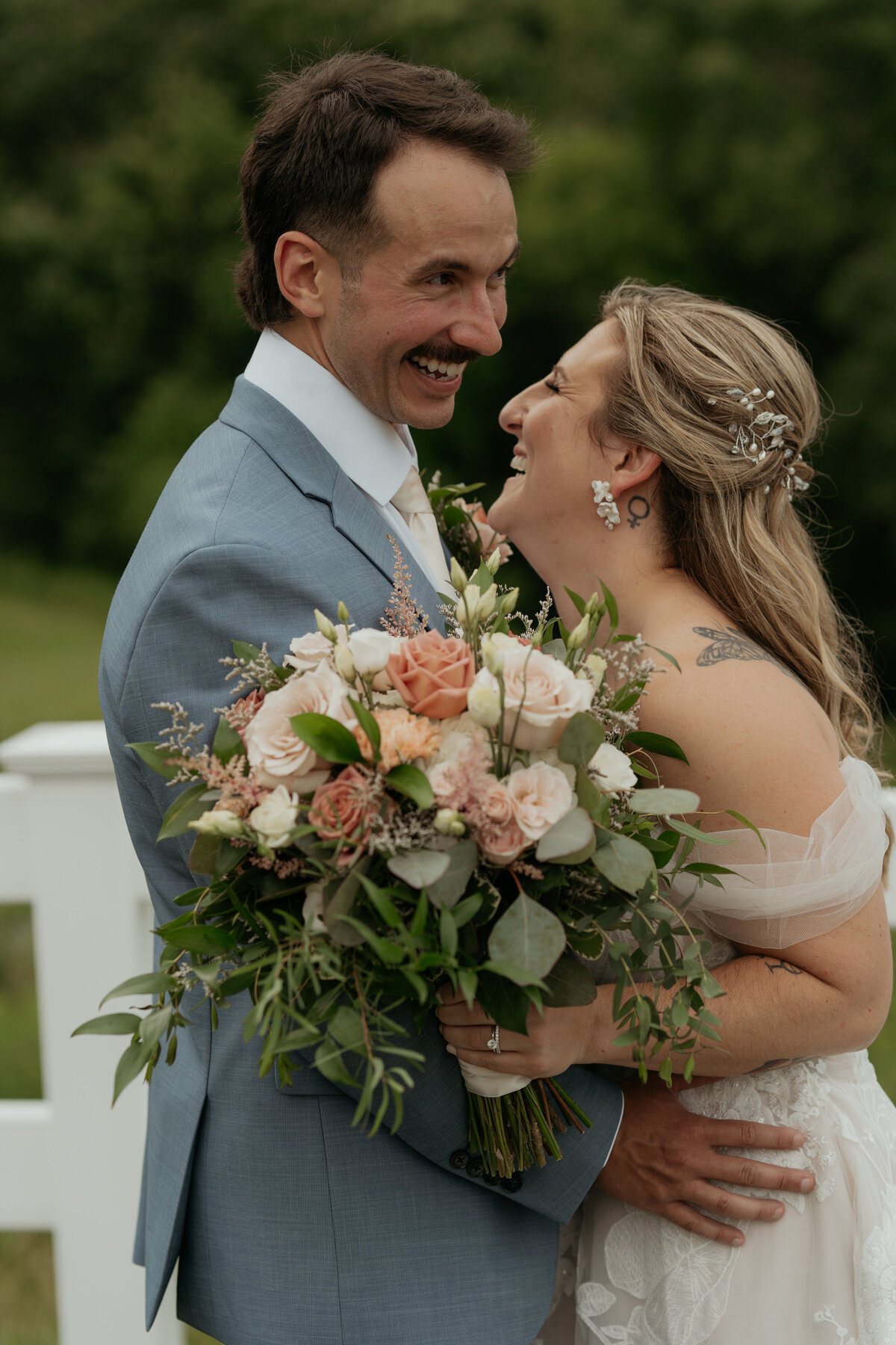 A joyful bride and groom share an intimate moment on their wedding day. The groom, wearing a light blue suit and white tie, smiles broadly while holding his bride. The bride, in an off-the-shoulder lace gown, beams with happiness and leans into her groom. She holds a large, lush bouquet of roses, peonies, and greenery. The couple is surrounded by a backdrop of greenery, emphasizing the natural and romantic atmosphere of their special day.