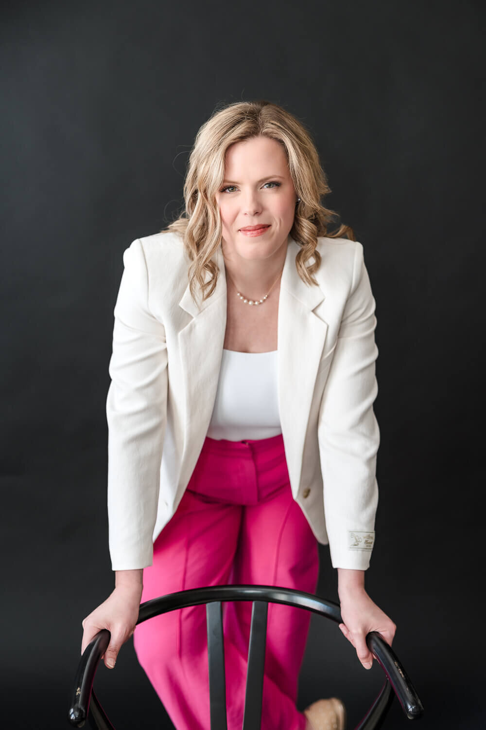 Confident female entrepreneur leaning on black chair wearing white blazer and pink trousers.