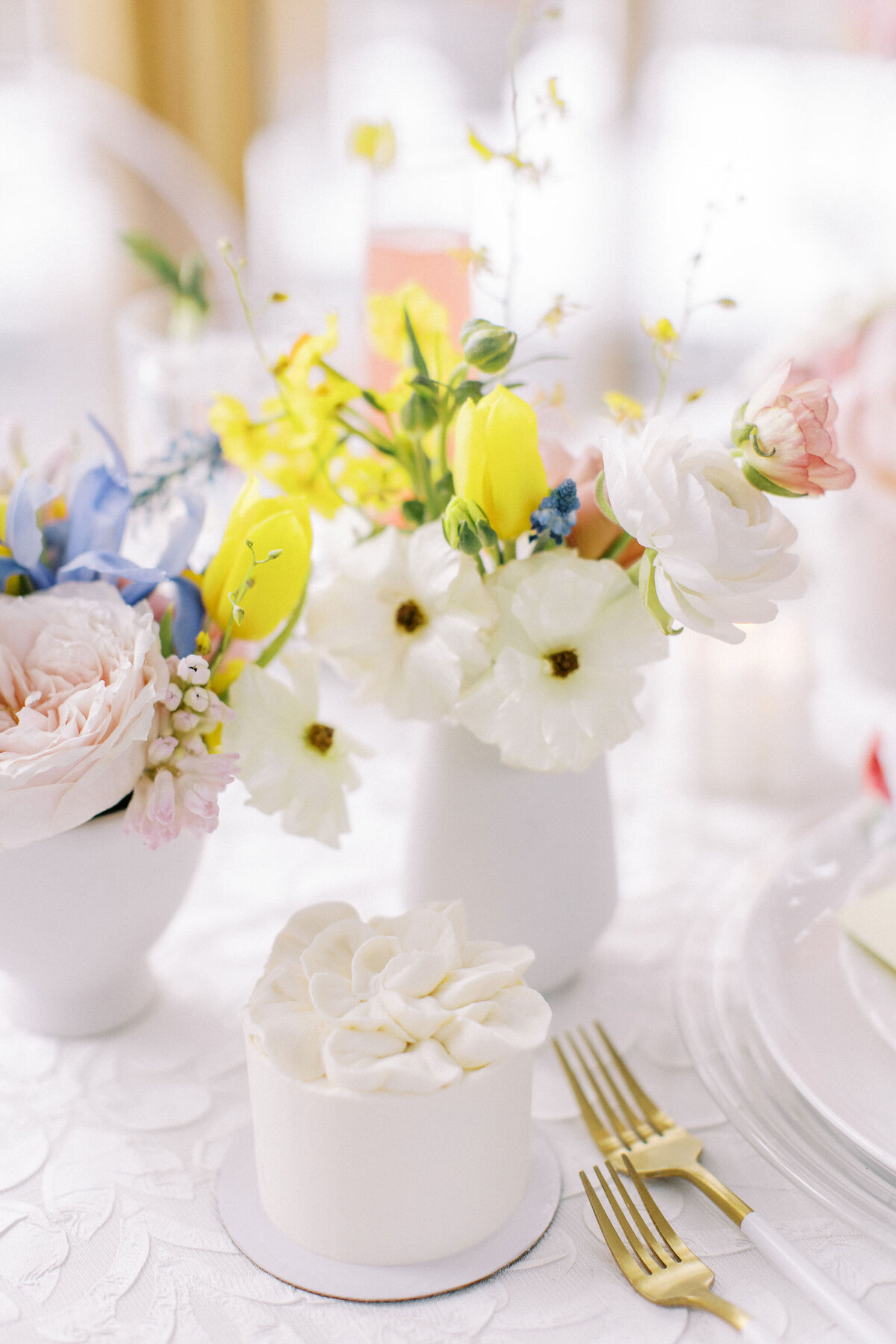 A small white cake with floral decoration is on a table next to white vases filled with yellow, blue, and white flowers. Two gold utensils are placed beside the cake, perfect for intimate weddings in Banff, Alberta.