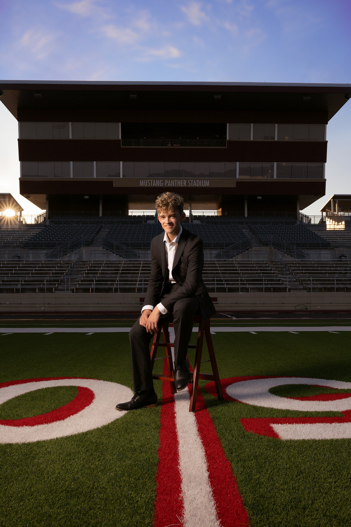 A high school senior poses confidently in a suit on a wooden stool at Mustang Panther Stadium for his senior photoshoot. The vibrant red and white lines on the football field complement his sharp look, capturing a bold moment in Dallas senior photography.