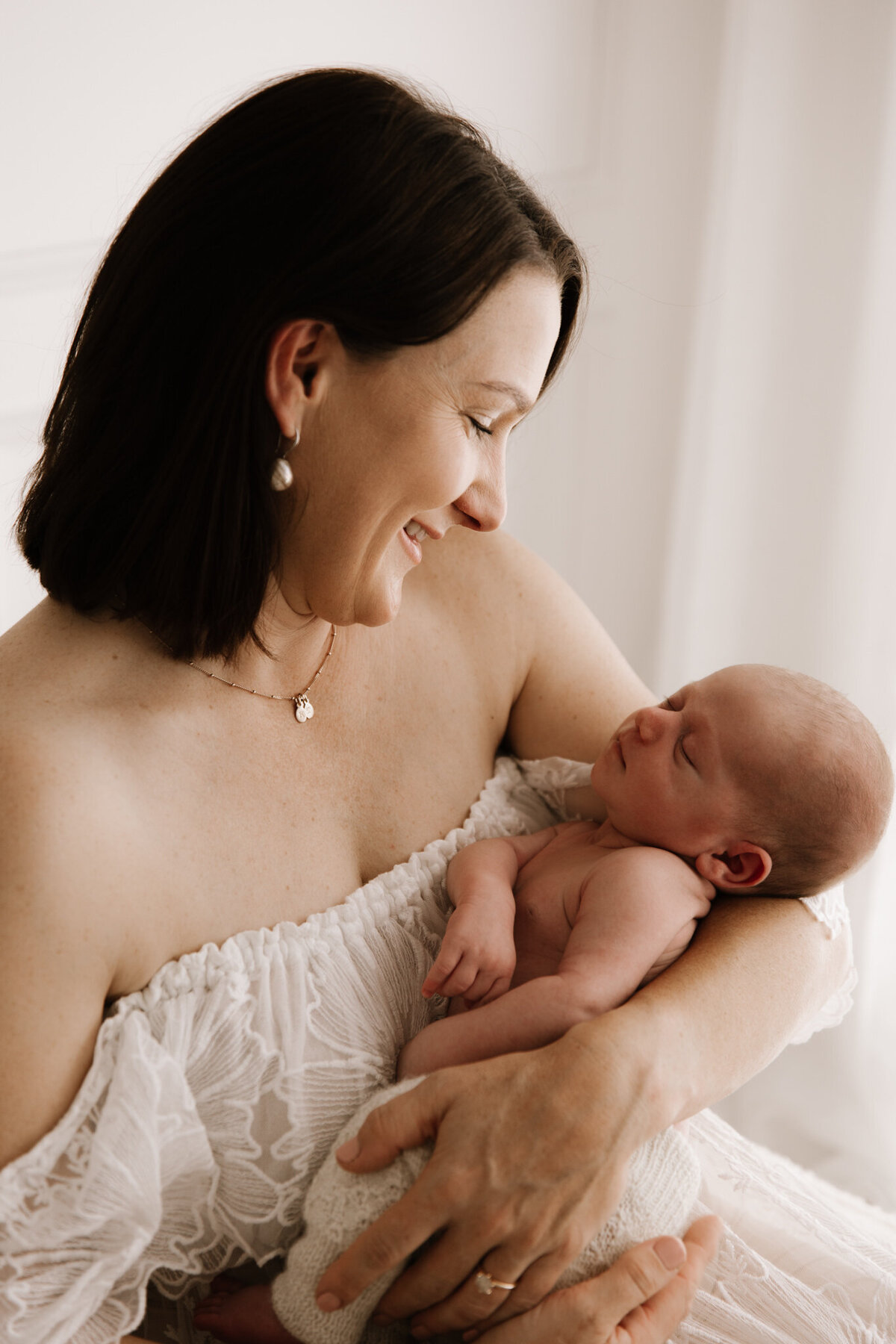 Close-up of a smiling mother in a white lace gown gently holding her sleeping newborn in a bright, soft-lit photography studio in Southwest Sydney.