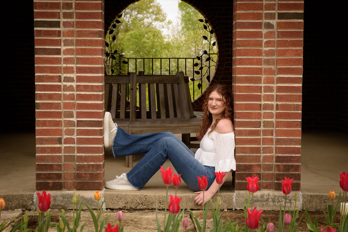 Green Bay East High School senior girl wearing a white sleeveless shirt and bell bottom jeans sitting behind a tulip garden in the red brick building at the Green Bay Botanical Gardens in Green Bay, Wisconsin.