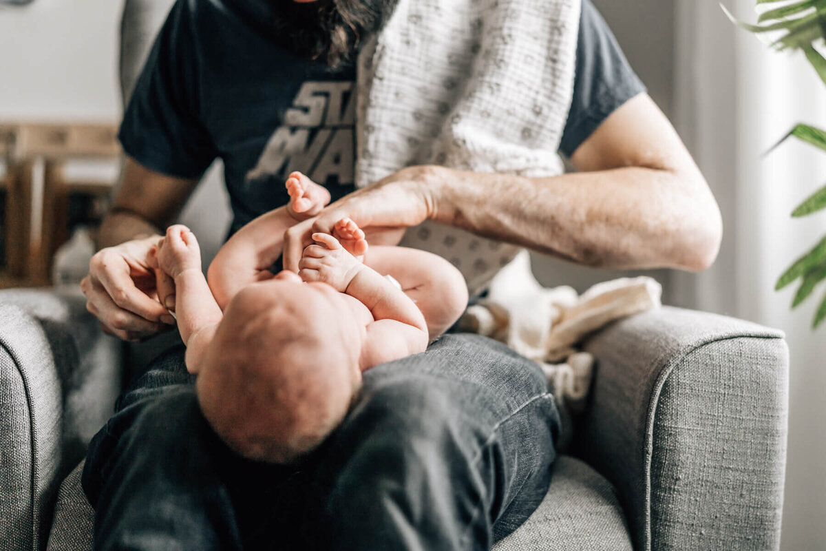 An image by Minneapolis newborn photographer Kate Simpson, of a newborn baby lying on her father's lap, having  her diaper changed, while Dad has tossed a burp cloth over his shoulder.