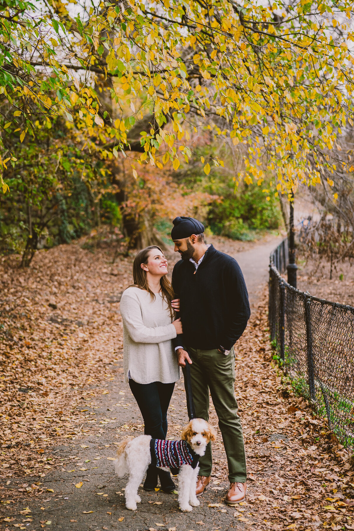 An engaged couple looking at each other at fort tryon park in nyc with their dog in the fall