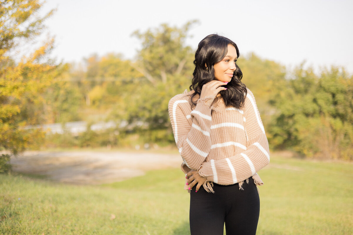 high school female senior, with long black hair, standing in grass with a tree line behind her, looking away from the camera smiling softly, wearing black leggings, and light tan and white stripped shirt