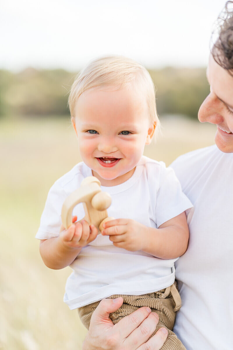 a toddler boy is held by his father and plays with a wooden toy car during their Austin family session.