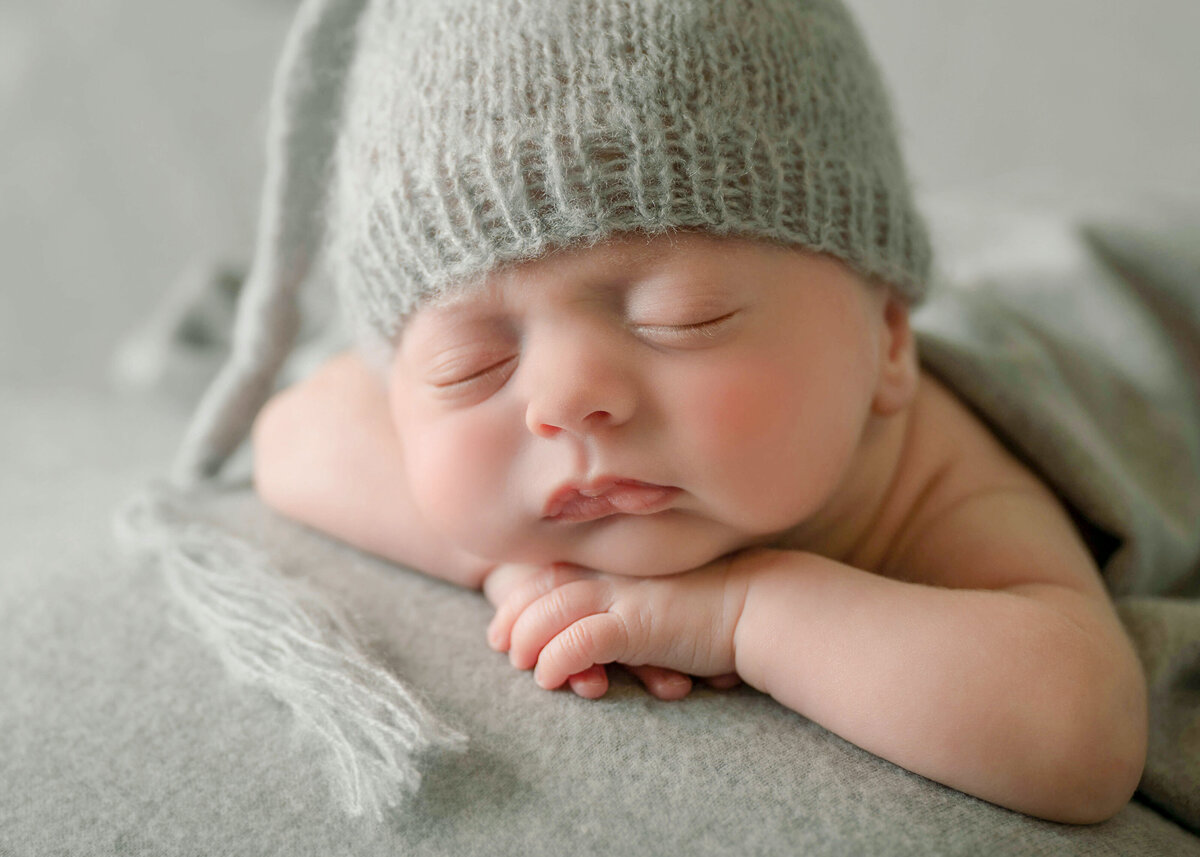 A rosy cheeked newborn baby sleeping on his tummy with his chin on his hands, on a gray background and wearing a grey  night cap.