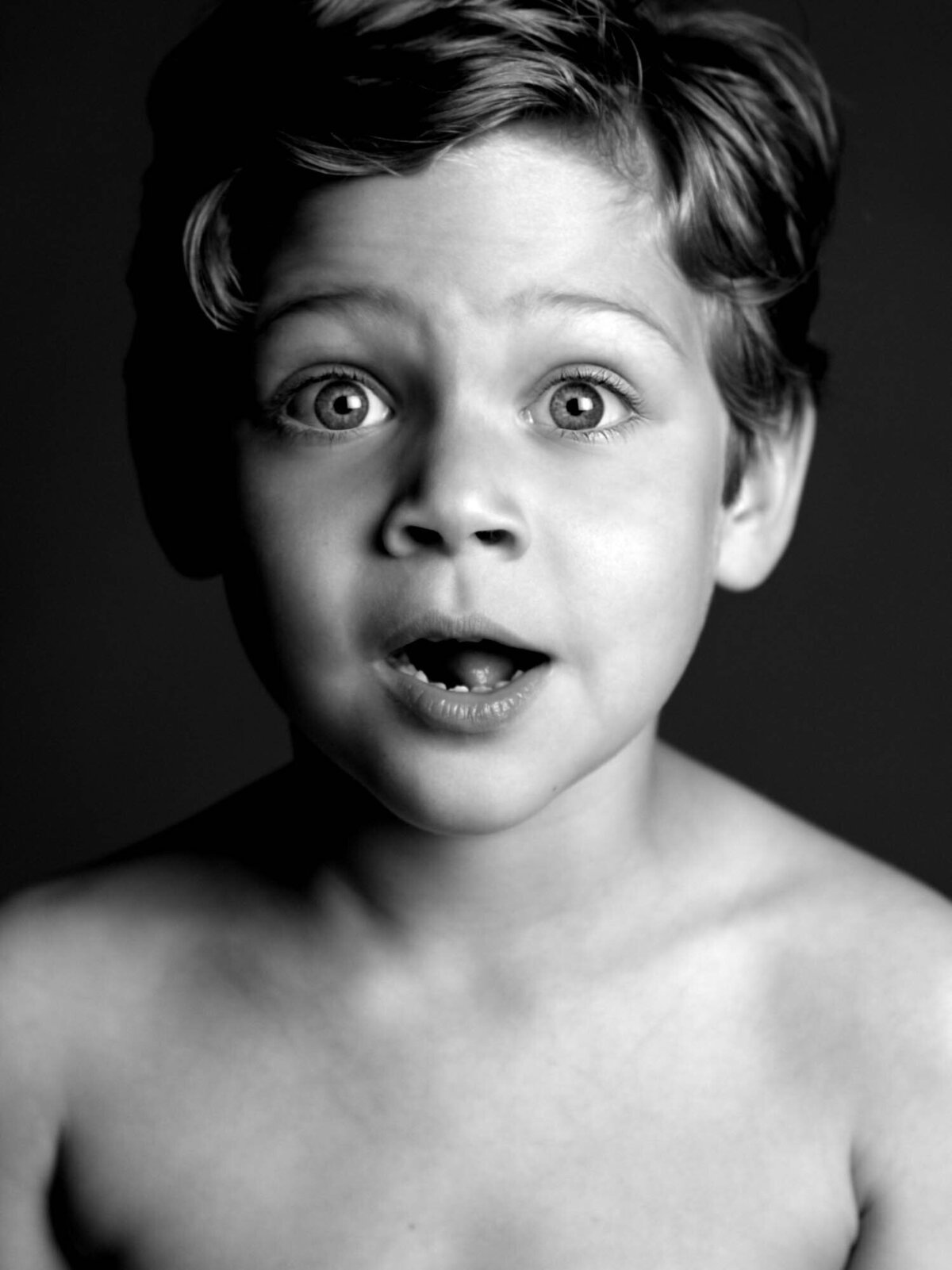 Black-and-white photo of a young child with wide eyes and open mouth, conveying surprise or awe. The background is plain and the child is shirtless.