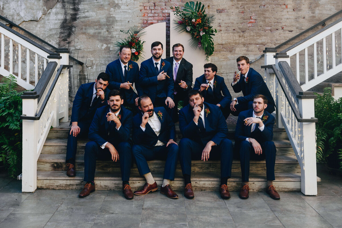 A group of ten men in blue suits pose playfully on wooden stairs, some crouching and some sitting. Behind them is a brick wall with a green and orange floral arrangement.