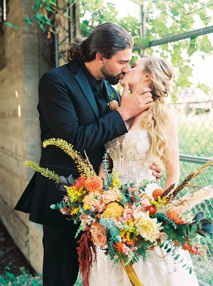 Bride and groom wearing a white wedding gown and black tuxedo kiss in front of a fence with large bouquet of flowers in hand.