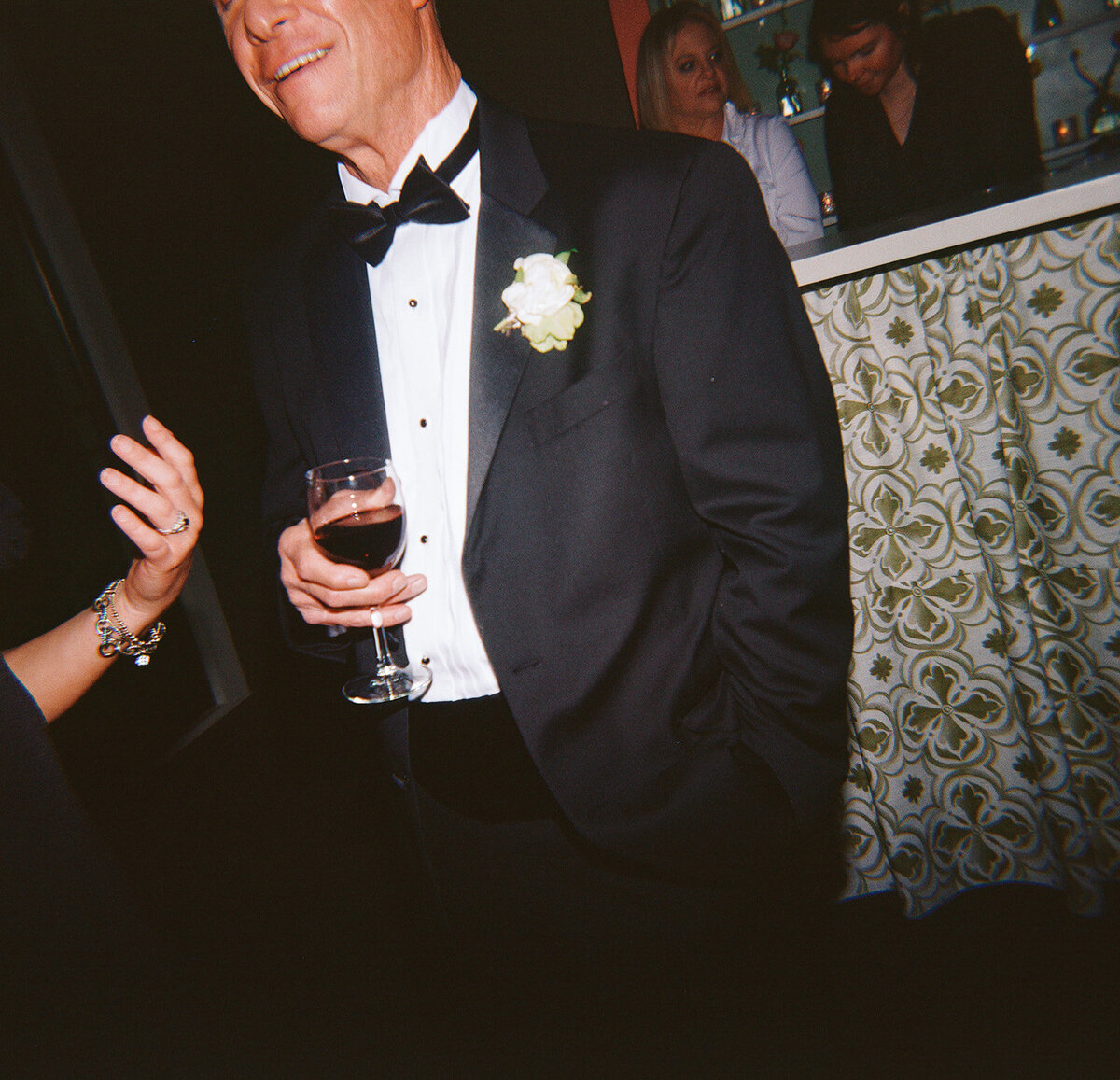 Close-up of a wedding guest in a black tuxedo holding a glass of red wine, with a white boutonniere pinned to his lapel, during the reception at Burritt on the Mountain.