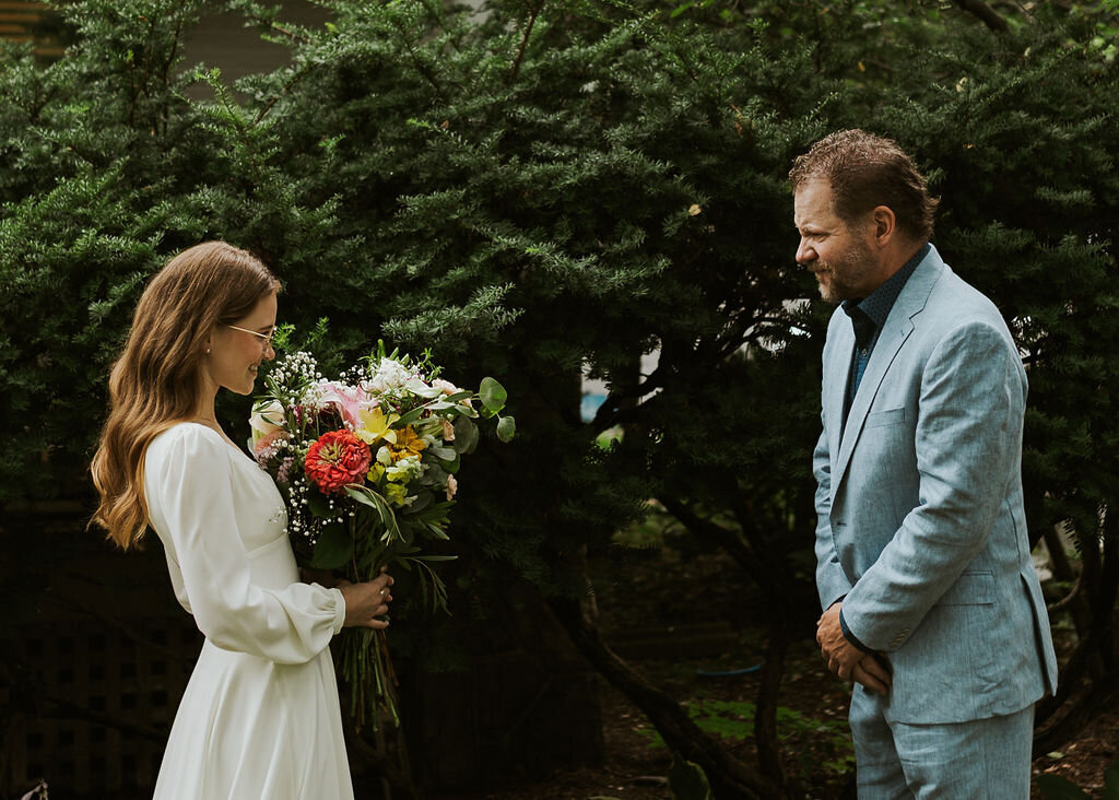 Bride holding a colorful bouquet stands siideways facing father wearing all blue suit. They are looking sweetly at eachother.