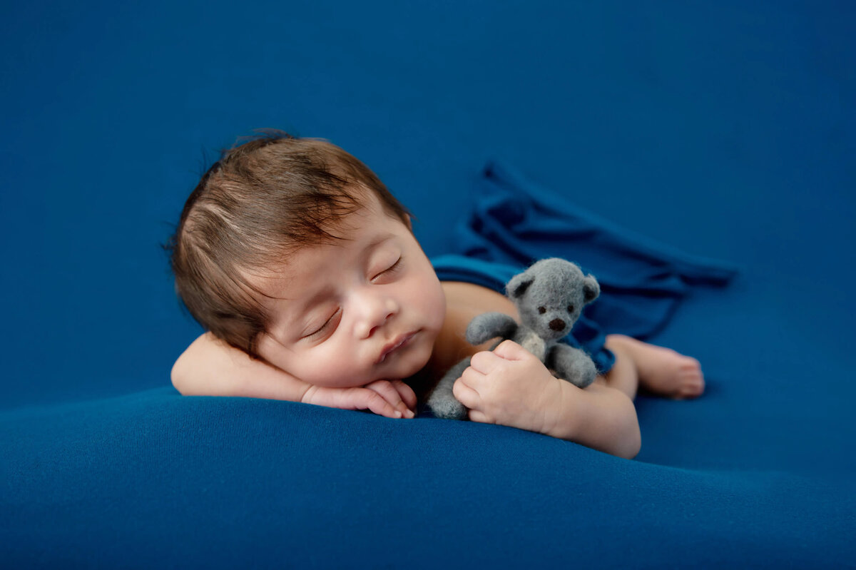 newborn boy laying on a blue background holding a blue teddybear for his newborn photos in Burlington