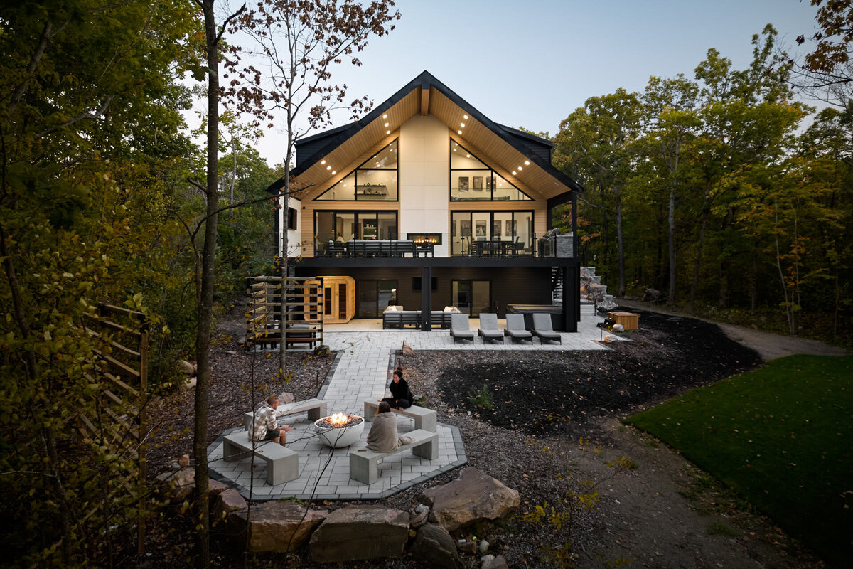 Aerial drone view of Ecobay Chalet in Port Severn, Muskoka Region, highlighting a luxurious chalet-style home ideal for upscale cottage rentals. The property features expansive glass windows, a glowing firepit seating area, a sauna, and a modern outdoor living space, surrounded by lush forest. Captured by a drone architectural photographer specializing in luxury properties in cottage regions.