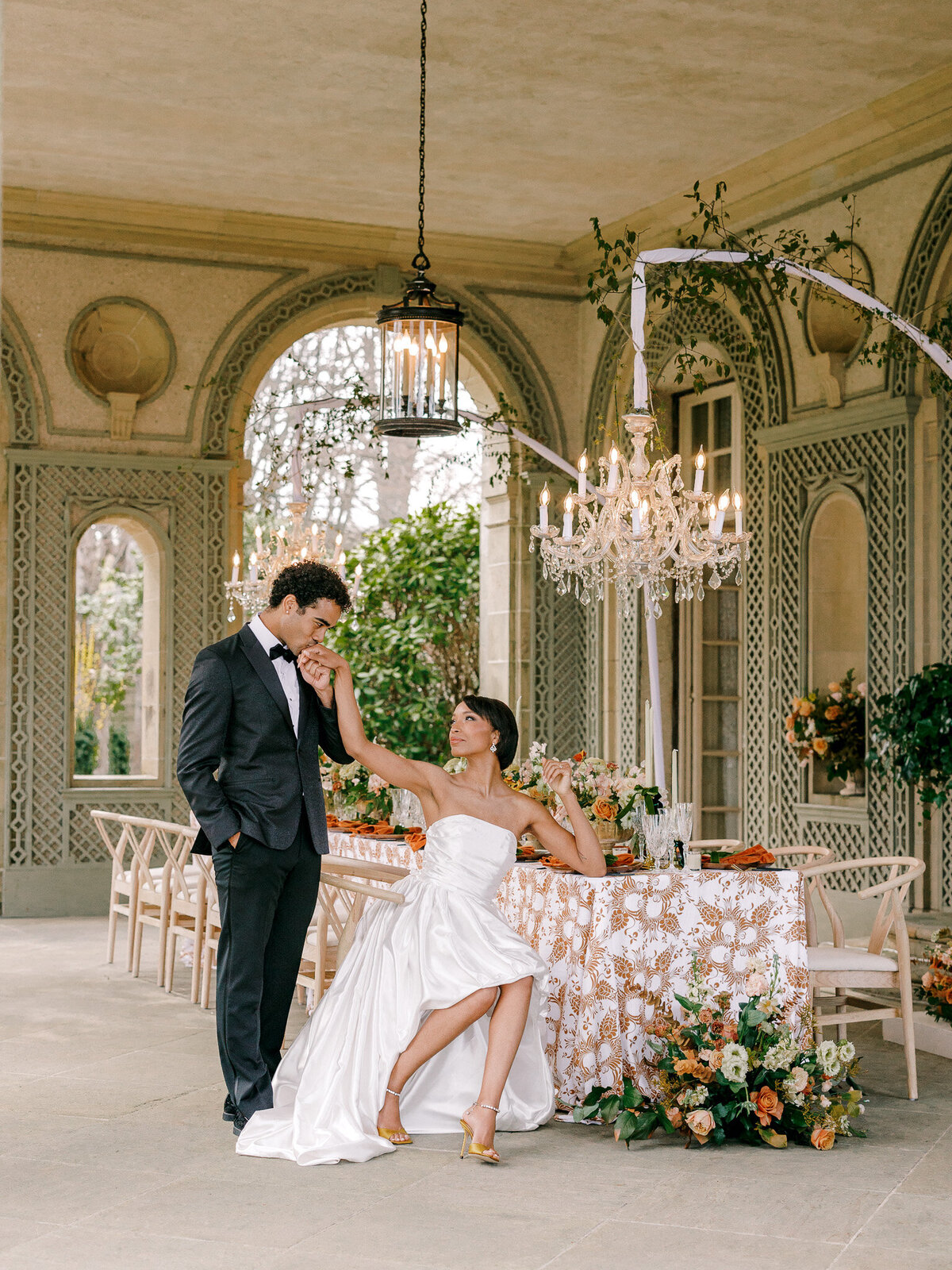 BIPOC groom stands while kissing the hand of his bride who is seated at wedding reception table